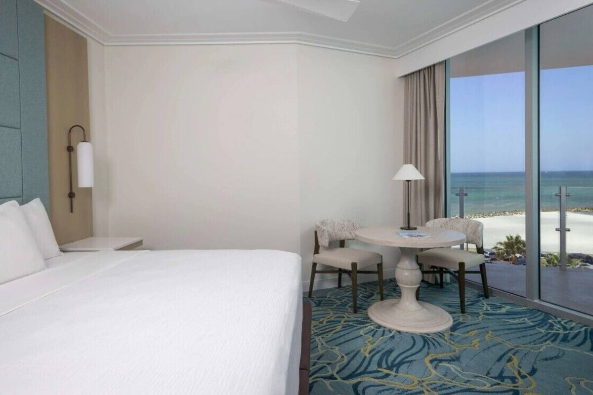 A hotel room with a large bed, a dining table with chairs, and a view of the beach.
