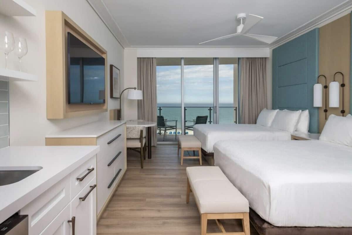 A hotel room with two beds, a desk, and a balcony with a view of the sea.