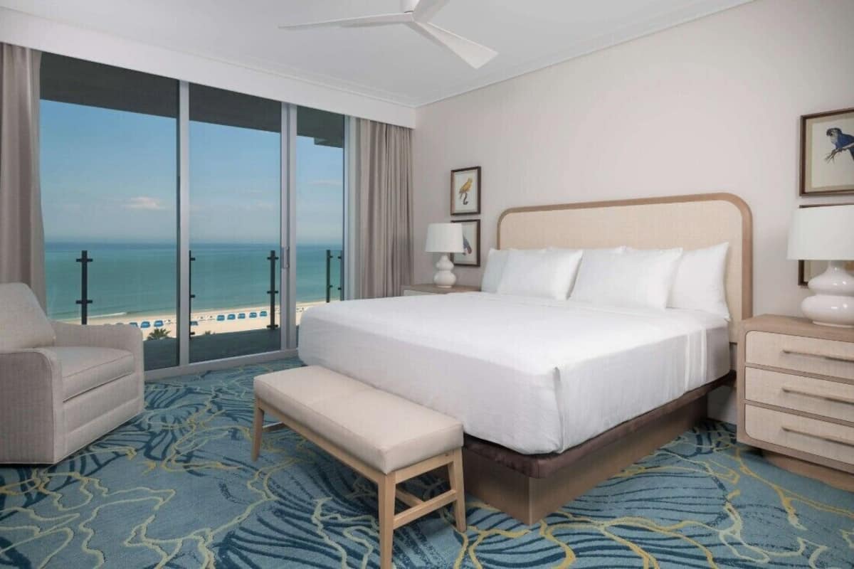 A hotel room with a large bed, a bench, a desk, and a view of the ocean.