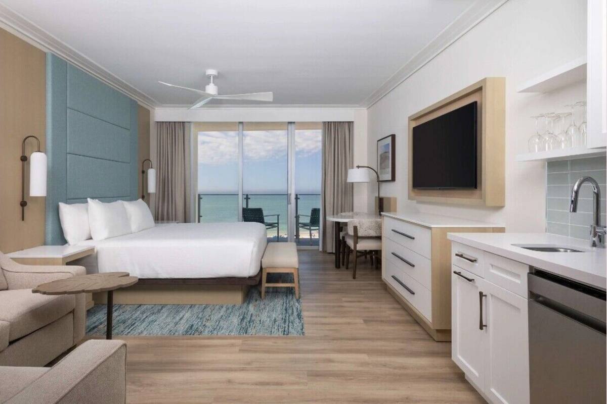A modern hotel room with a large bed, a desk, a kitchenette, and a balcony with a view of the ocean.