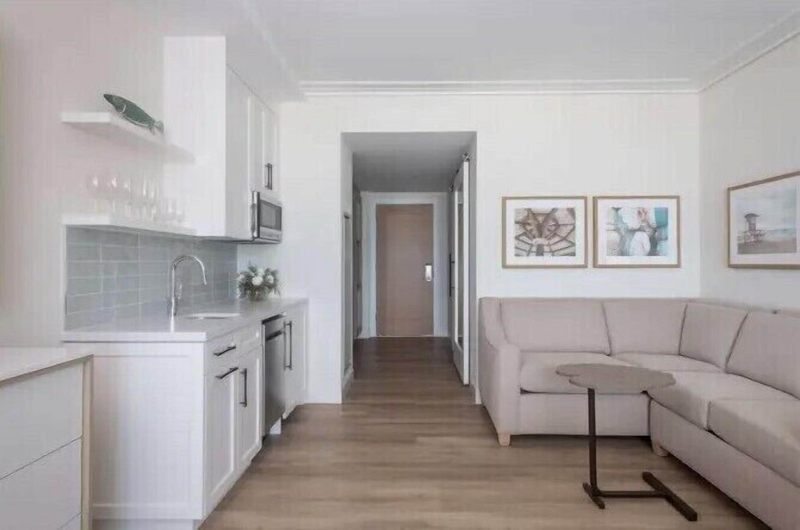 Eine moderne Küche mit weißen Schränken, einer Mikrowelle und einem Spülbecken. Im Wohnbereich befindet sich ein beiges Sofa und ein kleiner Tisch.