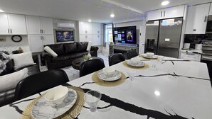 A modern living room with a black leather sofa, a dining table set with white plates and gold placemats, and a TV displaying various channels. - The Ardmore Executive Suite (Winston-Salem)