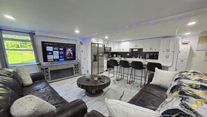 A modern living room with a large flat-screen TV, leather sofas, a coffee table, and a kitchen area with bar stools. - The Ardmore Executive Suite (Winston-Salem)