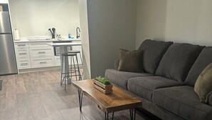 Un salon moderne avec un canapé gris, une table basse en bois et une cuisine en arrière-plan.