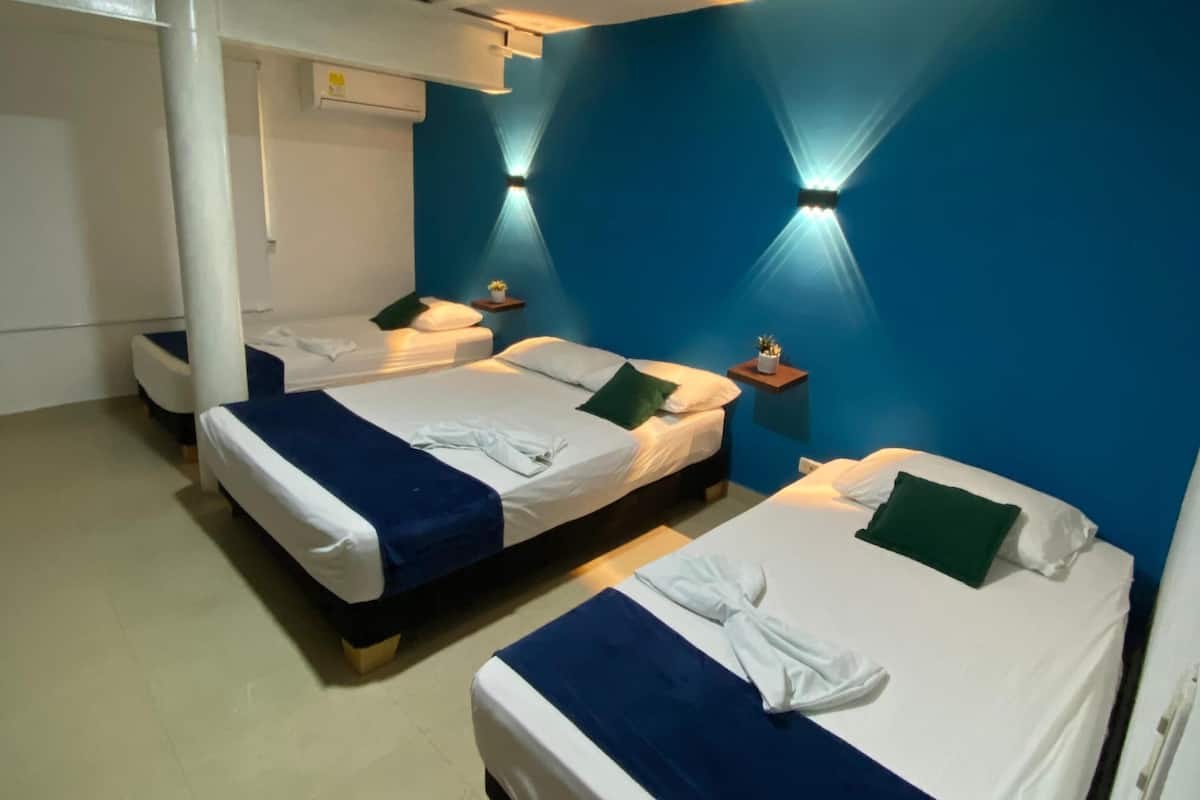 A hotel room with two beds, a white air conditioning unit, a small table with a plant, and a wall-mounted light fixture.