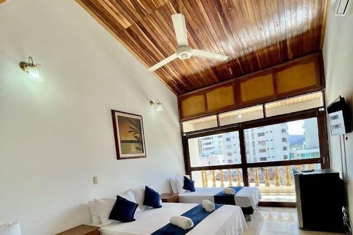 A hotel room with three beds, a ceiling fan, a large window with a view, and a desk.