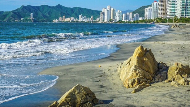 Uma praia com ondas, um primeiro plano rochoso e um horizonte urbano com edifícios.