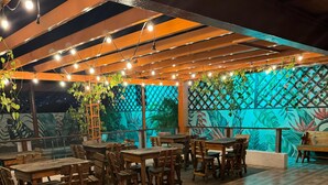 Uma área de refeições ao ar livre com mesas e cadeiras de madeira, luzes de fada e um mural com folhagem tropical.