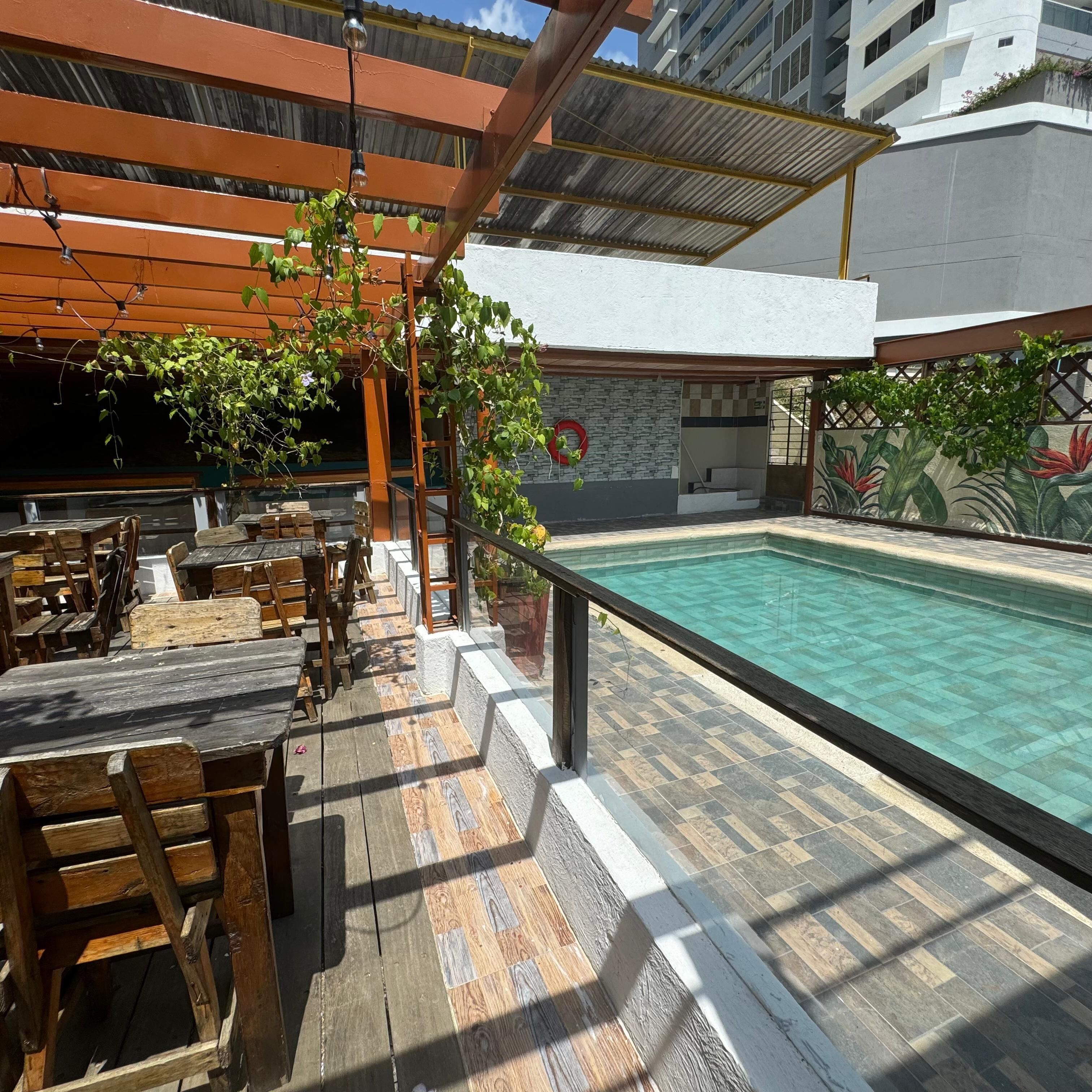 Ett utomhusområde med en pool, träbord och stolar, en pergola med klätterväxter och en stenmur med en väggmålning.