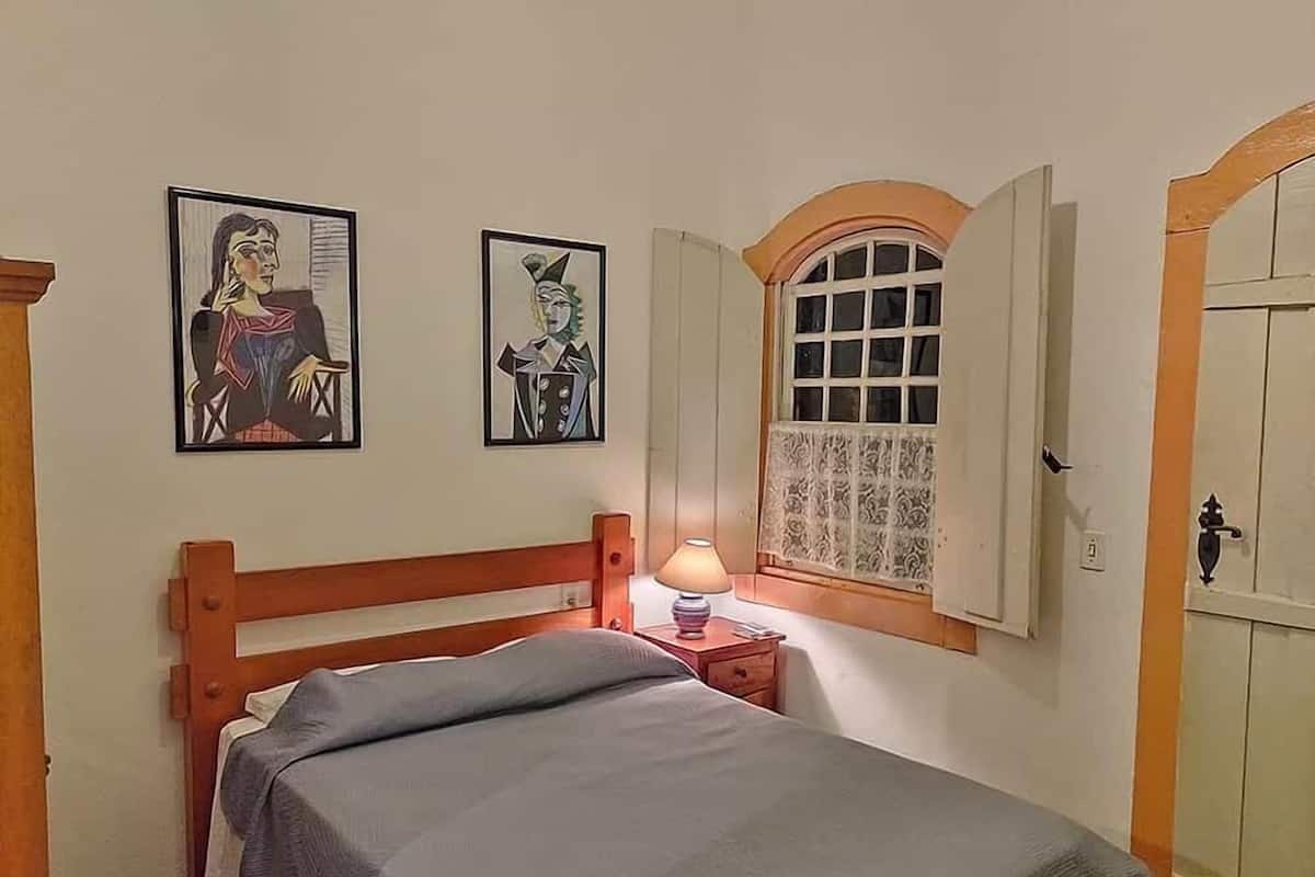 Un dormitorio con una cama de madera, una mesita de noche con lámpara, dos cuadros enmarcados en la pared y un ventanal de arco.