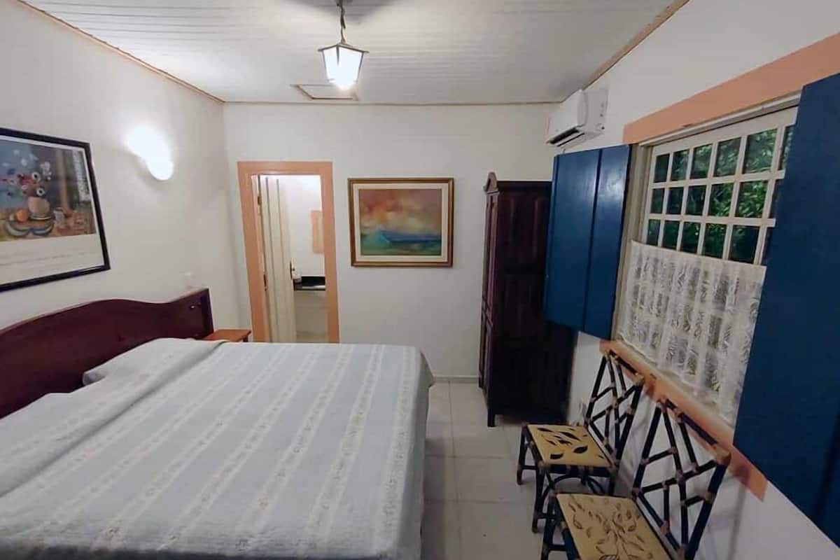 Un dormitorio con cama, escritorio y sillas, ventana con vistas, ventilador de techo y un cuadro en la pared.