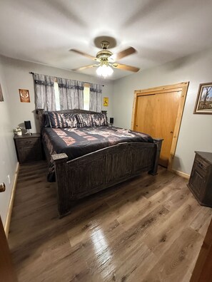 Un dormitorio con cama de madera, ventilador de techo, ventana con cortinas y puerta de madera.