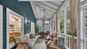 En overdækket veranda med sofa, stole, spisebord og et køkken, der kan ses igennem et vindue.