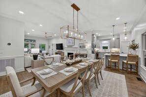 A modern kitchen with white cabinets, a dining area with a wooden table set for six, and a living room with a sofa and a TV. - 25 Atlantic Beach, Seaside Kiawah, Steps to Ocean, Pool Access! (Kiawah Island)