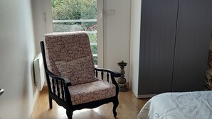 Um quarto com piso de madeira, uma poltrona estampada, uma cama com colcha estampada, um guarda-roupa e vista para o verde através de uma janela.