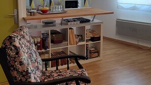 Sala de estar com piso de madeira, poltrona estampada, área de cozinha com micro-ondas e janela com persianas.