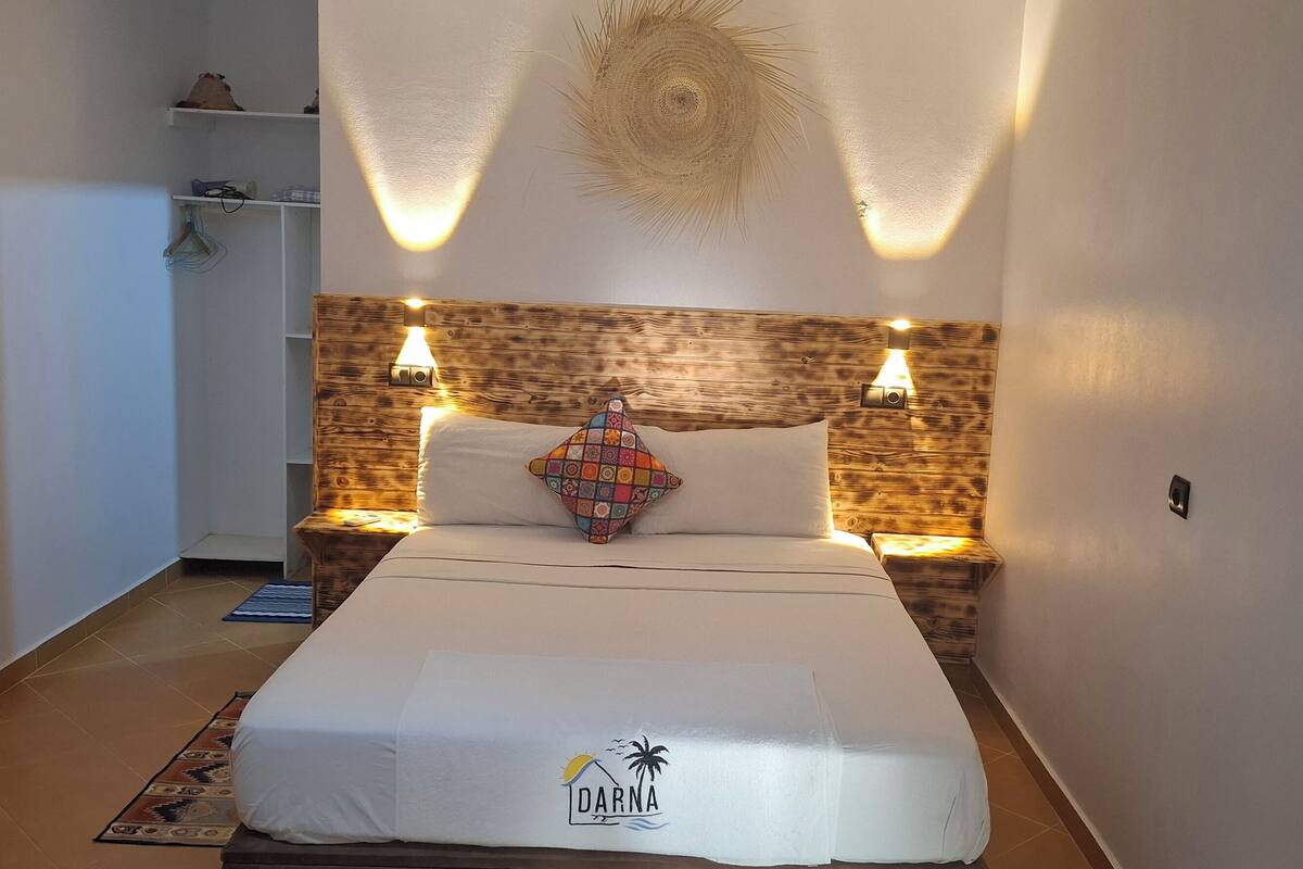 A neatly made bed with a white bedspread and a logo, two bedside lamps, a wooden headboard, and a decorative wall piece.
