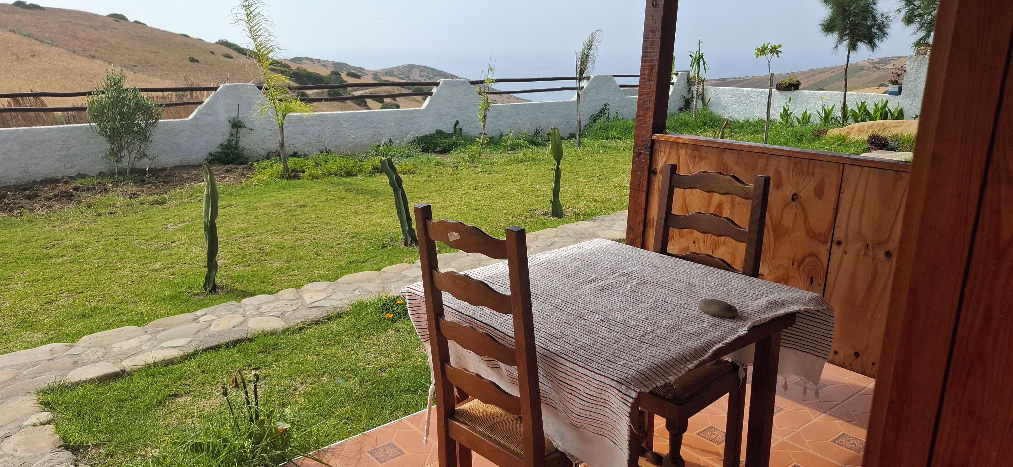 Un patio con una mesa y sillas de madera, un camino de piedra y vistas a un paisaje costero con un muro blanco y vegetación.