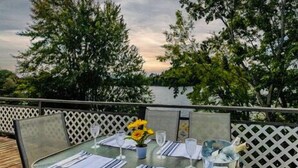 Un espace extérieur aménagé pour les repas, avec une table dressée pour deux personnes, entouré d’une clôture blanche en treillis, offrant une vue sur une étendue d’eau et des arbres.