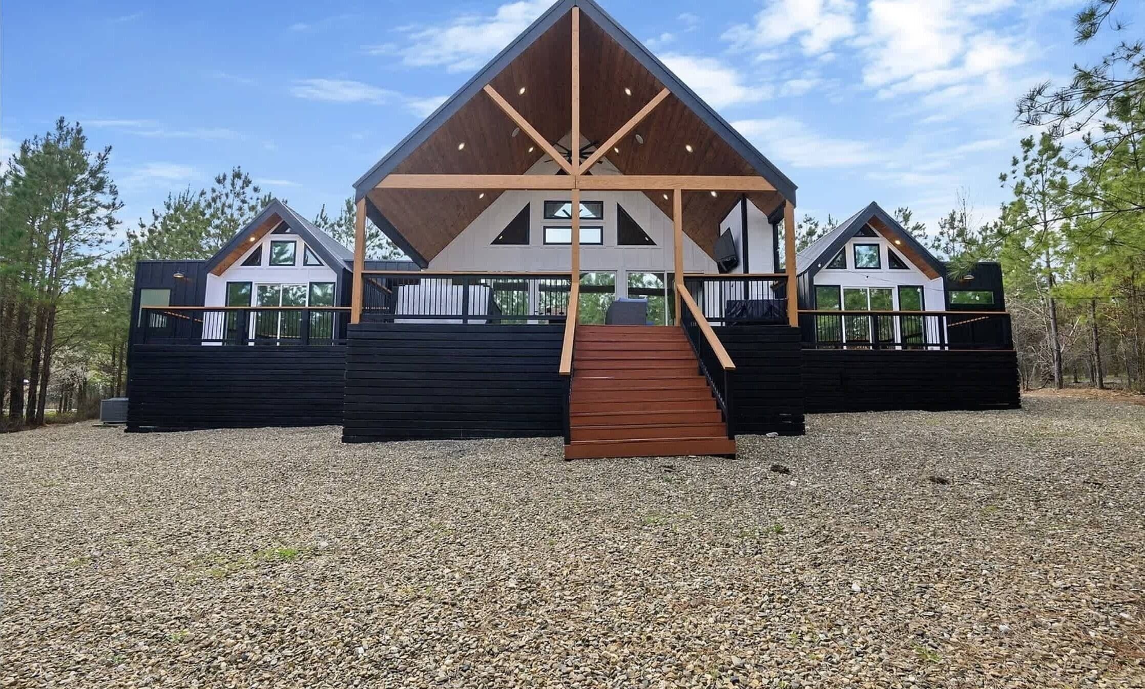 Een moderne chalet met een centraal houten gedeelte en twee zijvleugels, met een grindpad en omringd door bomen.