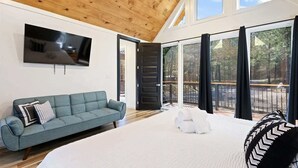 A modern bedroom with a large bed, a grey sofa, a TV mounted on the wall, and a balcony with a view of trees. - Couples - Family - Games - Hot tub - Sauna (Broken Bow)