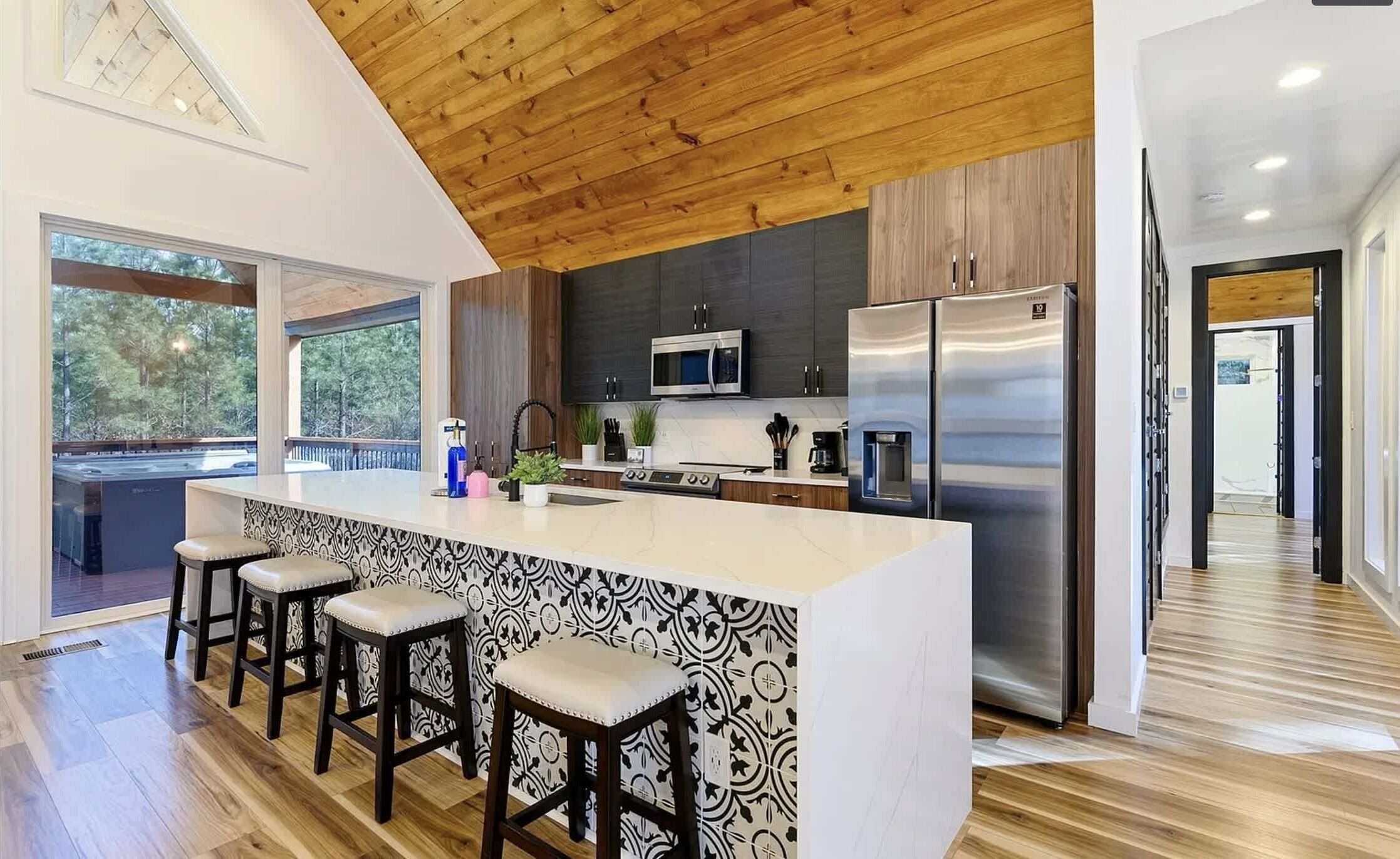 A modern kitchen with a wooden ceiling, a large island with bar stools, stainless steel appliances, and a view through the window.