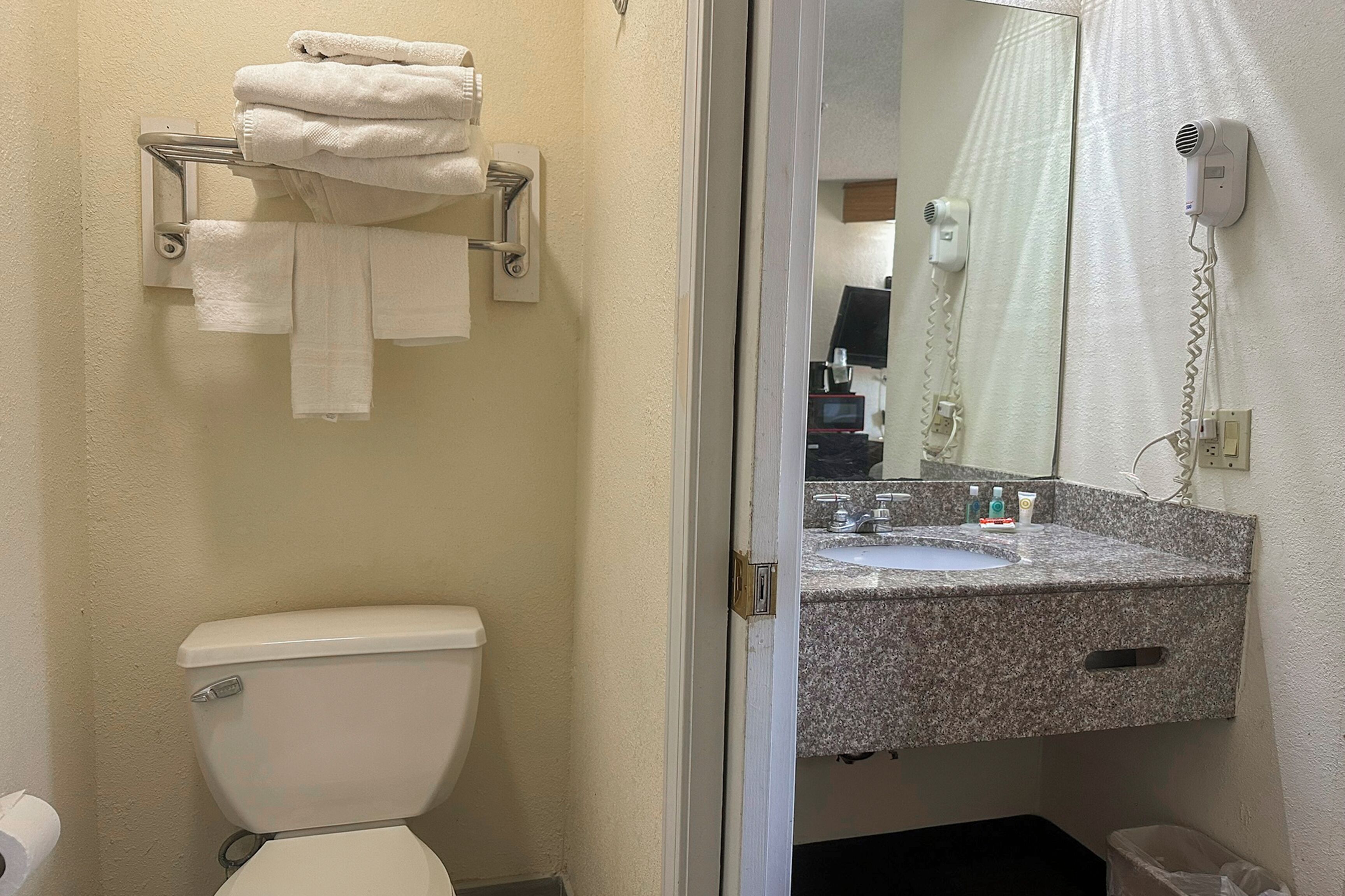 A bathroom with a toilet, a towel rack with folded towels, a sink with a mirror, and a hairdryer on the wall.