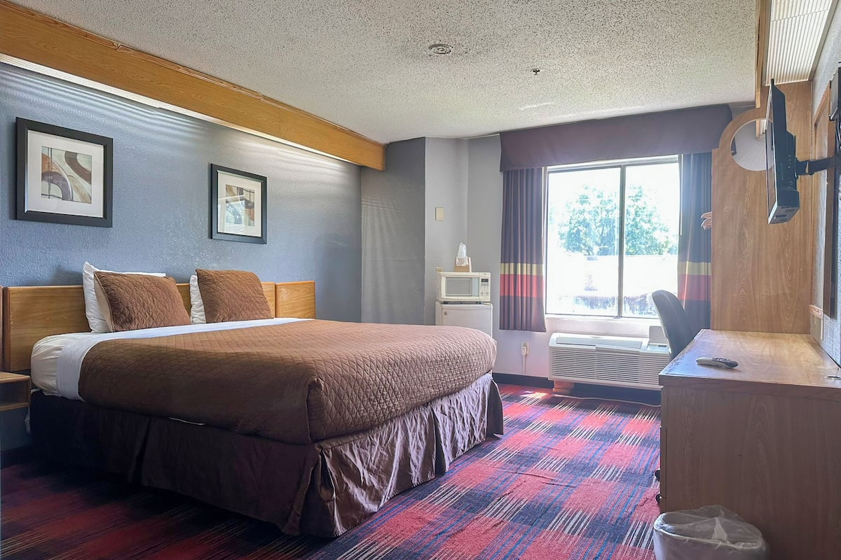 A hotel room with a large bed, a desk, a chair, a TV, and a window with curtains.