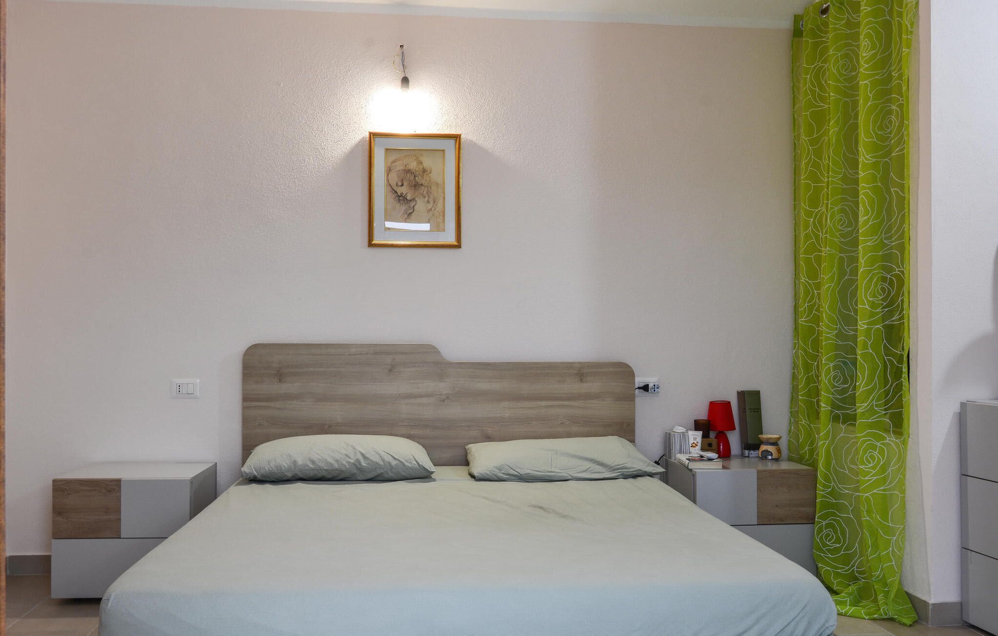 Ein Schlafzimmer mit einem Bett, zwei Nachttischen, einem Beistelltisch, einem gerahmten Bild an der Wand und einem grünen, gemusterten Vorhang.