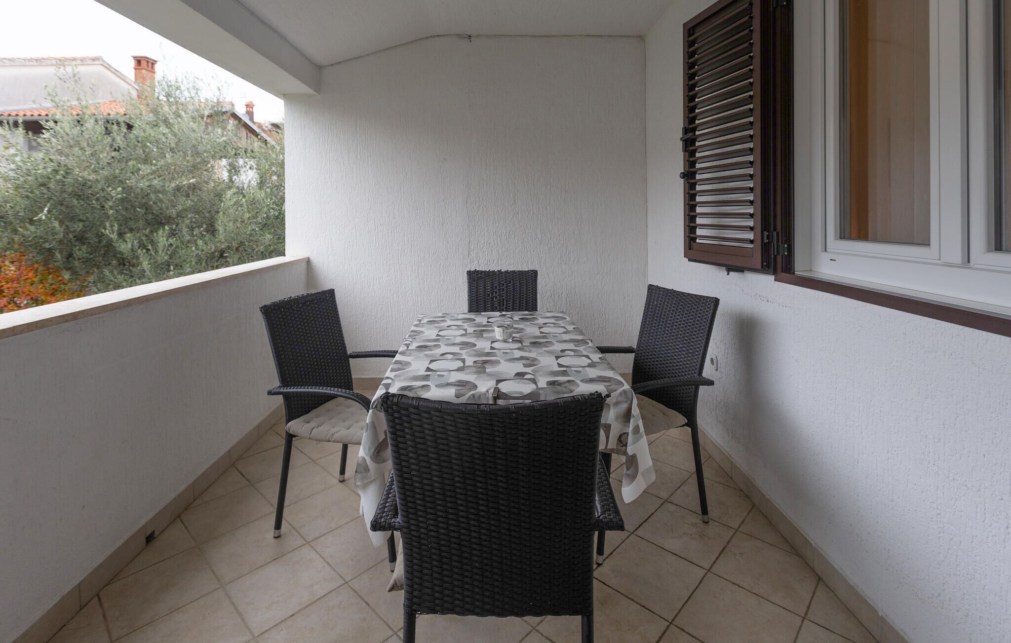 A balcony with a table and four chairs, a window with shutters, and a view of a building with a red roof.
