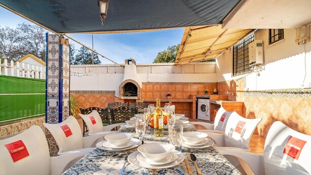 Un área de comedor al aire libre con una mesa dispuesta para seis personas, rodeada de sillas blancas con etiquetas rojas.