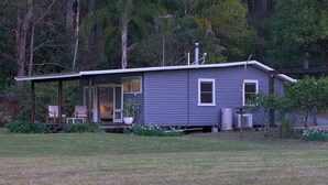 A small, single-story house with a porch, a covered outdoor seating area, and a garden with white flowers. - Kick back in a calm, stylish space in the foothills of the Watagan Mountains (Cooranbong)