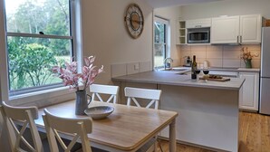 A modern kitchen with white cabinetry, a wooden dining table, and a vase of flowers. - Kick back in a calm, stylish space in the foothills of the Watagan Mountains (Cooranbong)
