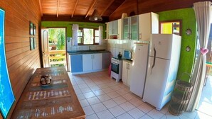 A kitchen with wooden cabinets, a white refrigerator, and a dining table with a basket of fruits. - Casa Vista Privilegiada - Piscina e Mar (Garopaba)