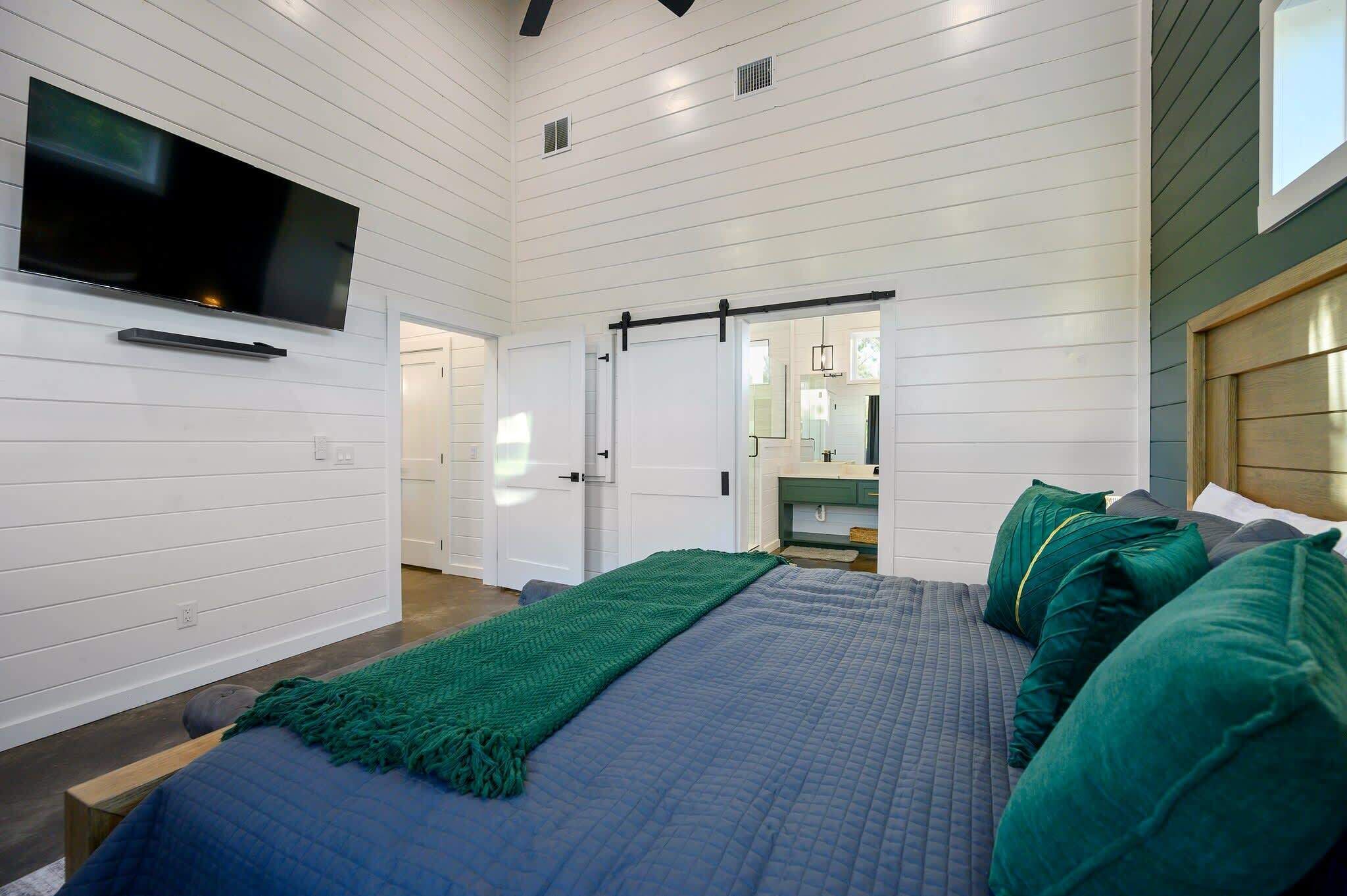 A bedroom with a large bed, a flat-screen TV mounted on the wall, and a sliding door leading to another room.