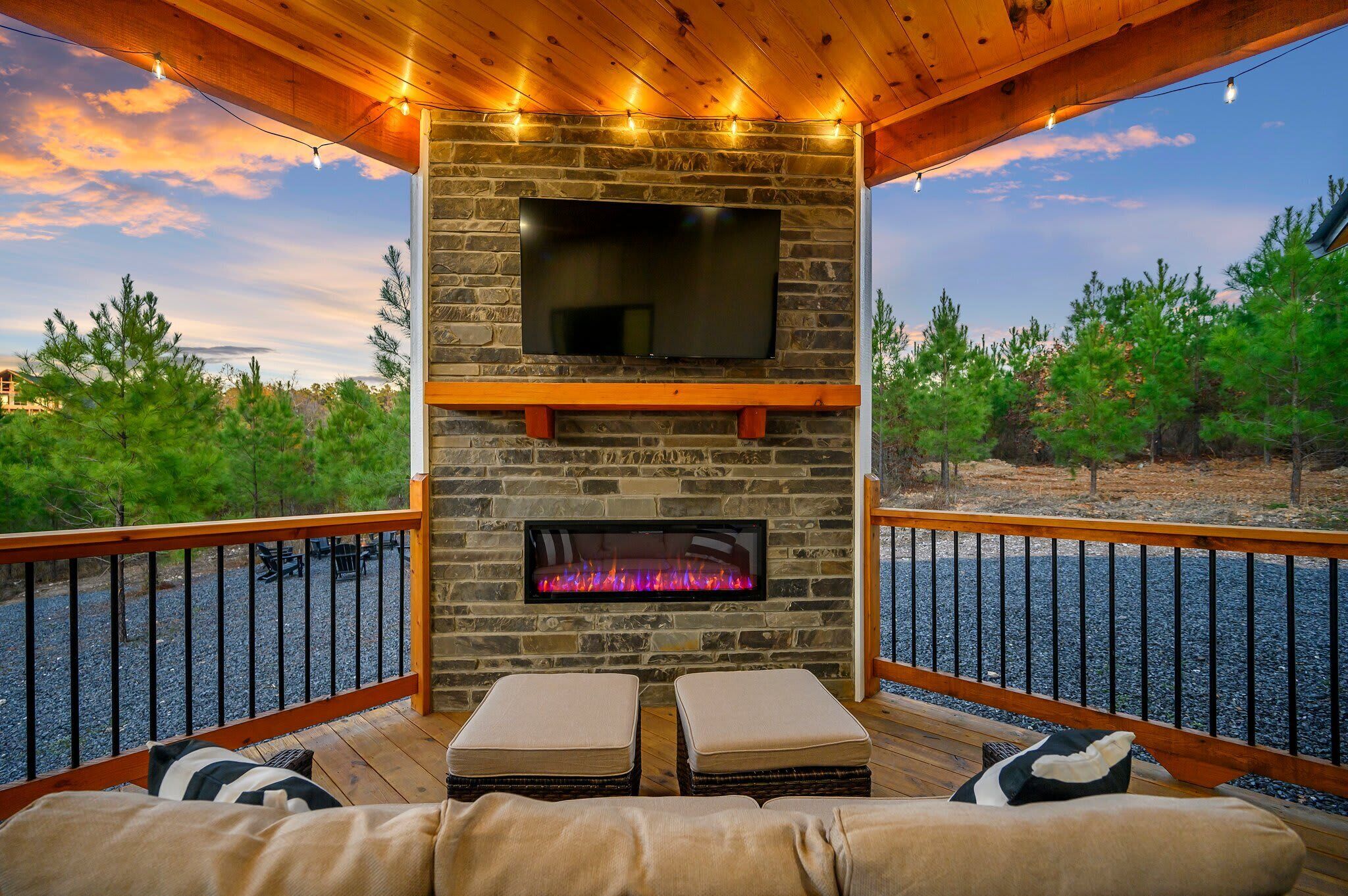 A stone fireplace with a flat-screen TV above it, wooden deck seating, and a view of trees and a sunset.