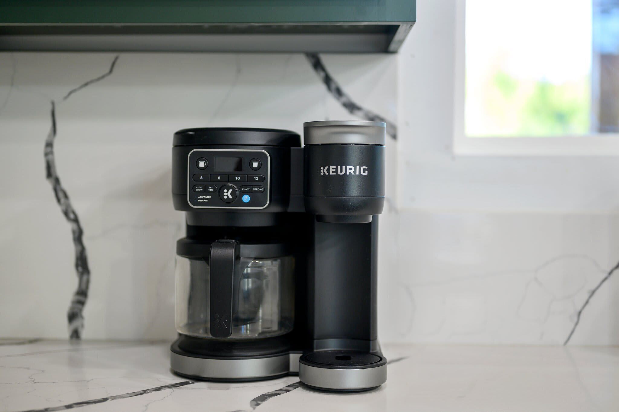 Eine Keurig-Kaffeemaschine mit Glasbehälter und Halterung für K-Cup-Kapseln auf einer Marmorarbeitsplatte.