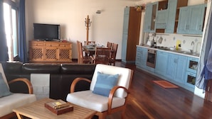 Sala de estar con un sofá, una mesa de centro de madera y una zona de comedor con mesa y sillas. Se observa una cocina con armarios azules y una estufa.