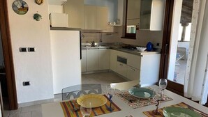 Una cocina compacta con armarios blancos, un pequeño comedor con platos y vasos, y una ventana con cortinas.