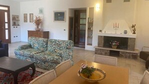 Una sala de estar con sofá, una mesa de comedor con un florero, una chimenea y una puerta que da a otra habitación.