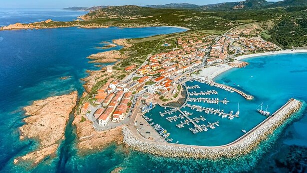 Vista aérea de un pueblo costero con un puerto deportivo, terreno rocoso y aguas azules cristalinas.