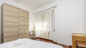 A bedroom with a bed, a wardrobe, a chair, a window with curtains, and a radiator.