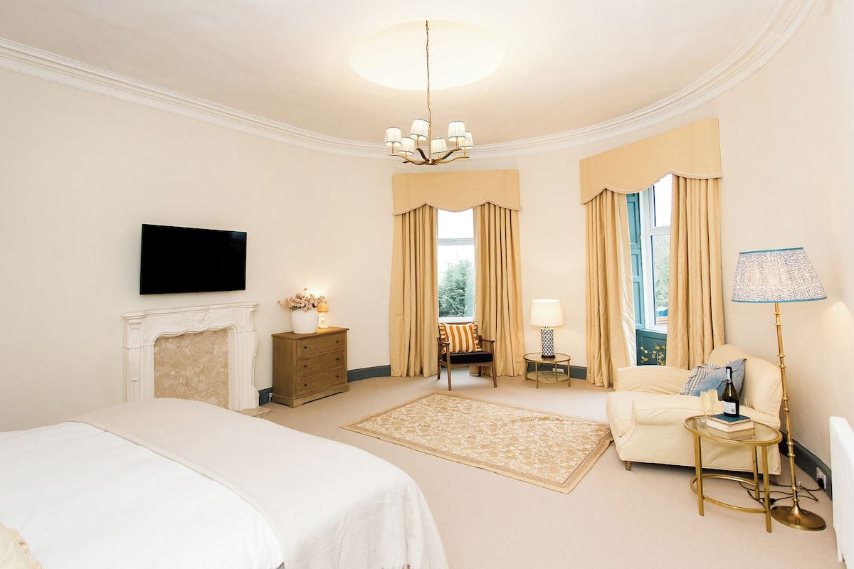 A bedroom with a large bed, a television, a fireplace, a chair, a table with a lamp, and a window with curtains.