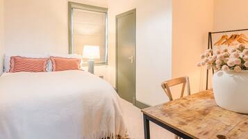 Habitación de hotel con cama, una mesa con un florero, una silla, una lámpara y una puerta.