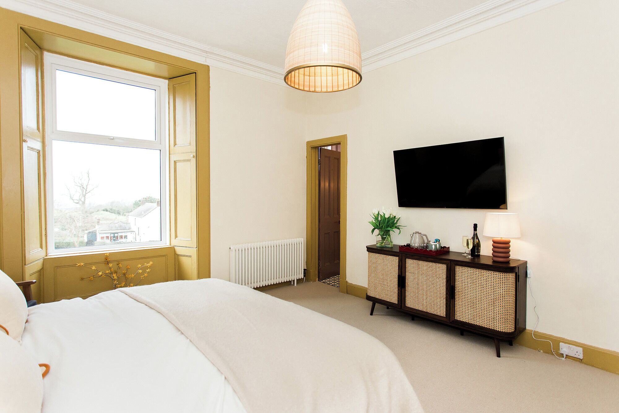 Habitación de hotel con una cama grande, televisión montada en la pared, mesita de noche con lámpara y ventana con vistas al exterior.