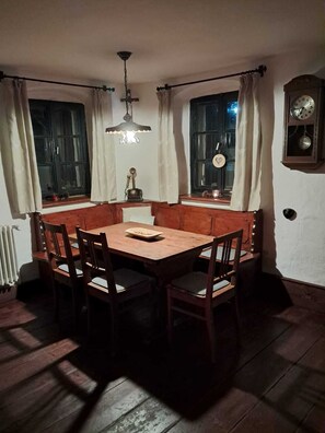 A dining room with a wooden table and chairs, two windows with curtains, a chandelier, and a wall clock.