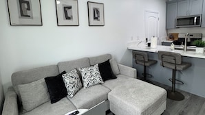 Un salon moderne avec un canapé d’angle gris, une petite table blanche sur laquelle se trouve un ordinateur portable, et un coin cuisine doté de meubles gris et d’un four à micro-ondes.