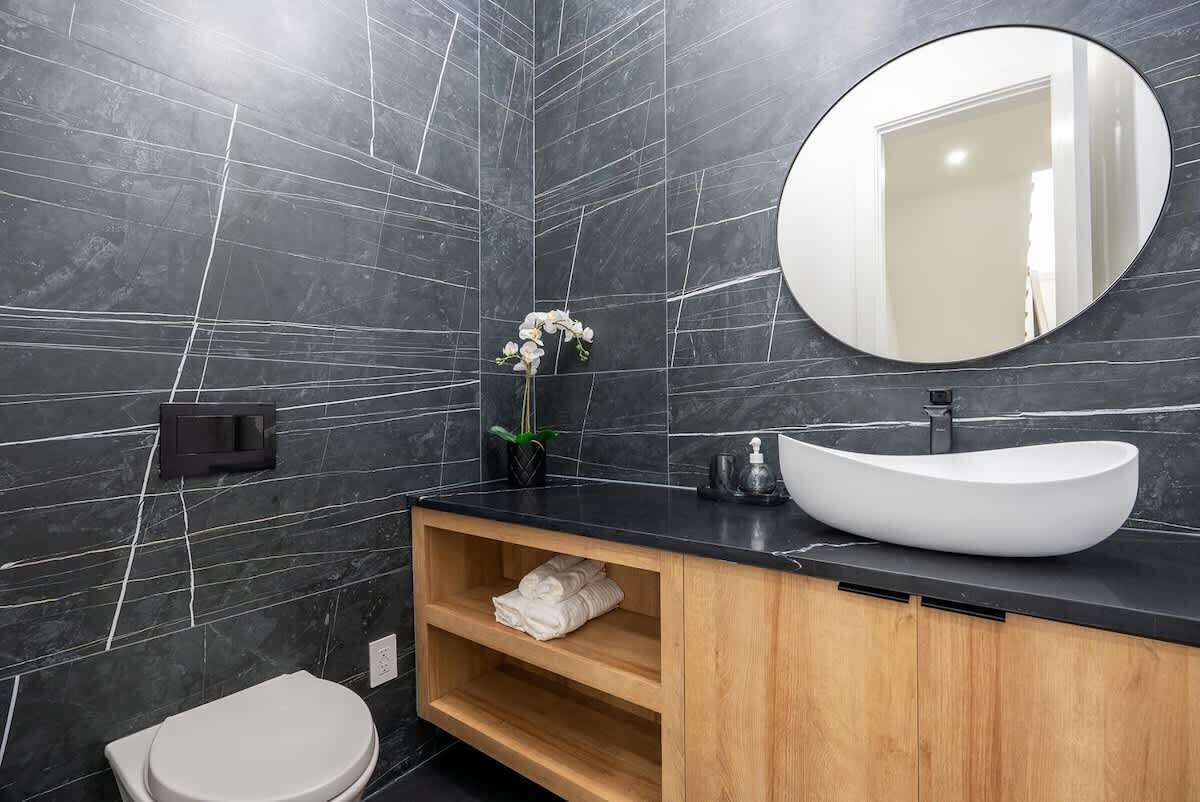 A modern bathroom with a black marble wall, a white sink, a round mirror, and a toilet.