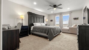 Ein Schlafzimmer mit einem Bett, Nachttischlampen, Deckenventilator, Kommode, Stuhl und einem Fenster mit Vorhängen.