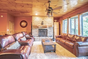 A cozy living room with leather sofas, a stone fireplace, and large windows.